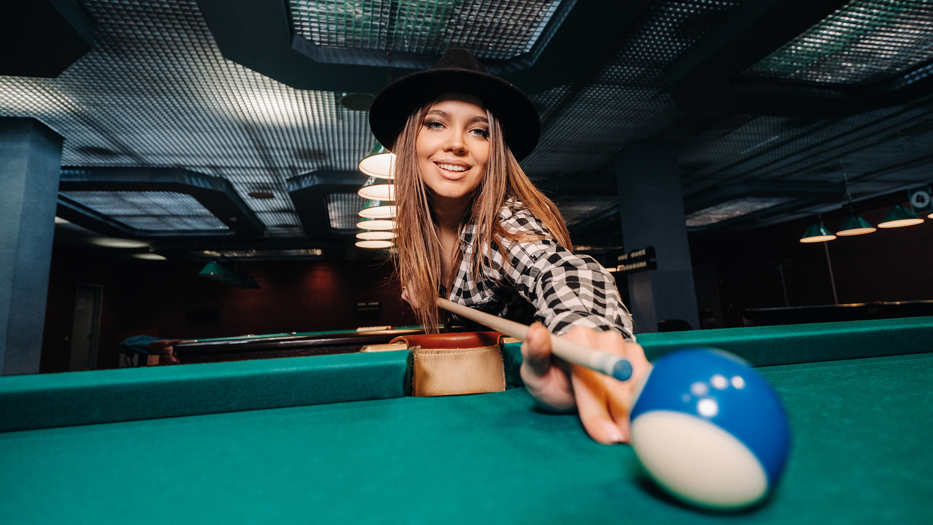 A girl in a hat in a billiard club with a cue in her hands hits a ball.Playing pool
